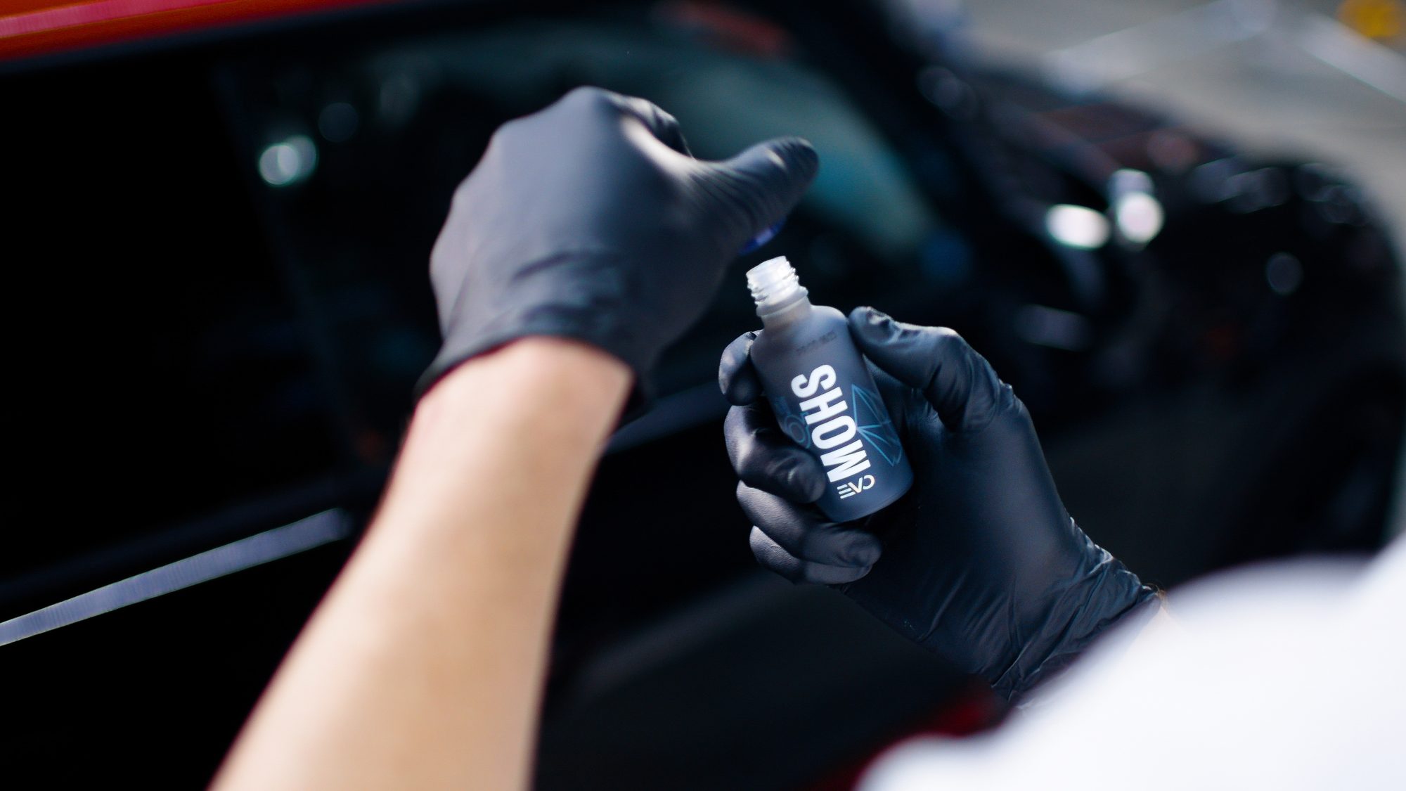 Geöffnete Flasche GYEON-Keramikversiegelung in der Hand, im Hintergrund dunkler Sportwagen